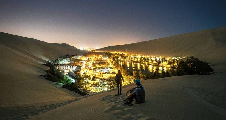 Vista nocturna de un valle con un pueblo resplandeciente y dunas de arena.