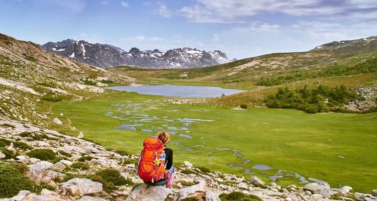 Trekker en solitario con mochila naranja descansa mirando un prado alpino verde y un lago rodeados de picos nevados