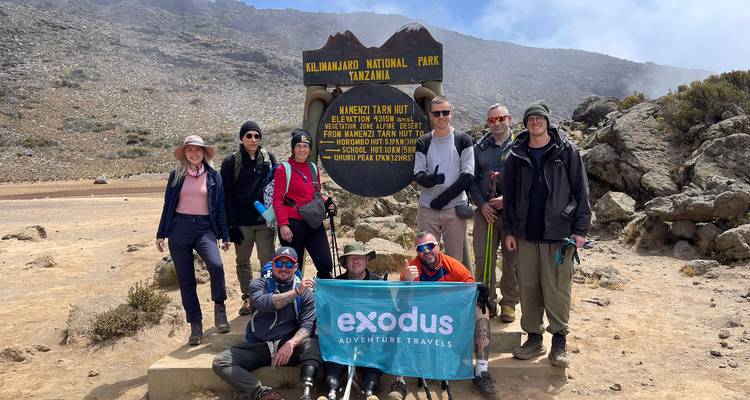 Groep trekkingstochten poseren bij het bordje van Mawenzi Tarn Hut terwijl ze een expeditievaan vasthouden op de Kilimanjaro.