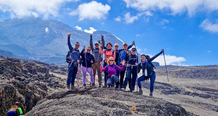 Negen vrolijke bergbeklimmers poseren op een rotsachtige uitsteksel met trekkingstokken omhoog geheven, de Mount Kilimanjaro rijzend tegen een levendige blauwe lucht achter hen.