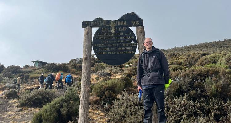 Een wandelaar staat glimlachend naast een houten Kilimanjaro National Park-wegwijzer omgeven door heide terwijl andere trekkersachter voorbijgaan.