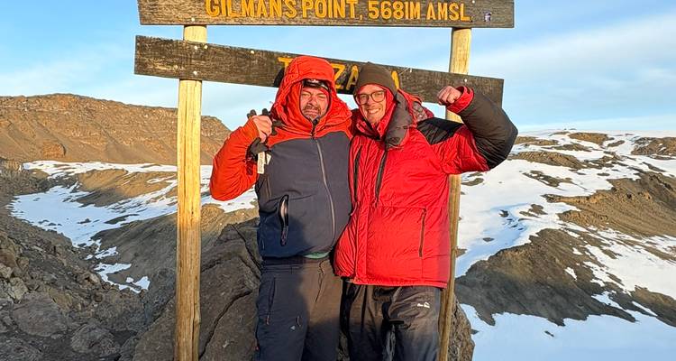 Twee trekkingstokers in warme jassen vieren het uit onder het houten bord van Gillman's Point 5681 m met overal sneeuw en vulkanisch terrein om hen heen.