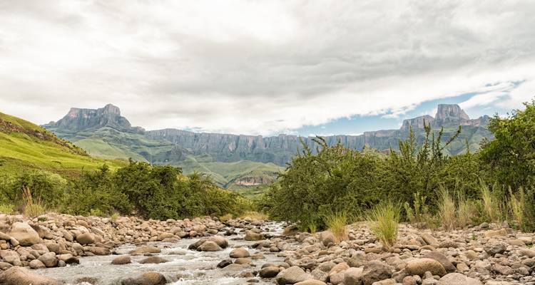 Rotsachtige beek stroomt door een groen dal onder de Drakensberg-escarpment onder bewolkte lucht.