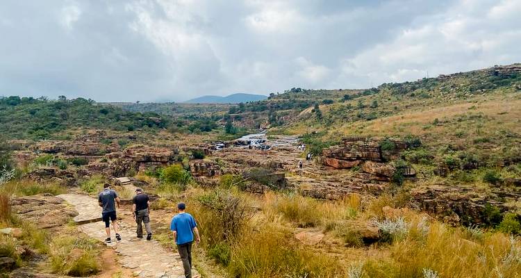 Reizigers wandelen langs een rotsachtig canyonpad onder bewolkte hemels met uitzichtpunten in de verte.