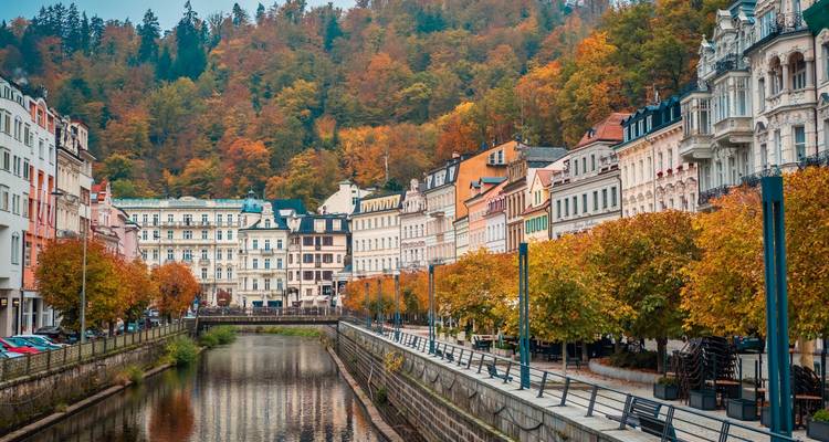 Kanaal geflankeerd door kleurrijke kuuroordgebouwen en gouden herfstbomen, met beboste heuvelrug op de achtergrond in Karlovy Vary.