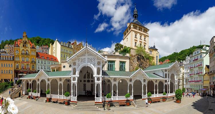 Brede hoekopname van Karlovy Vary's sierlijke witte Marktcolonnade omlijst door kleurrijke historische gevels and weelderige heuvels.