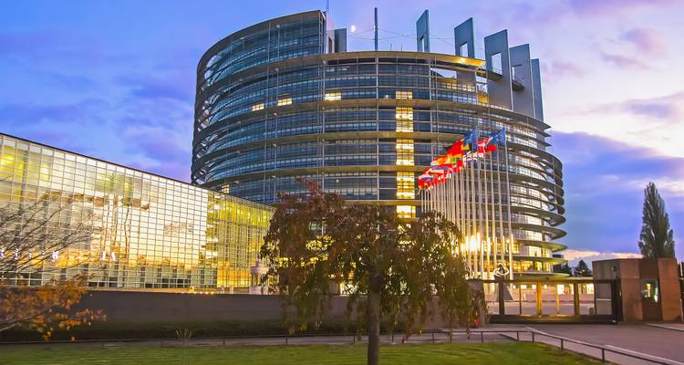 Bâtiment moderne en verre courbé du Parlement européen illuminé au crépuscule avec une rangée de drapeaux de l'UE qui flottent.