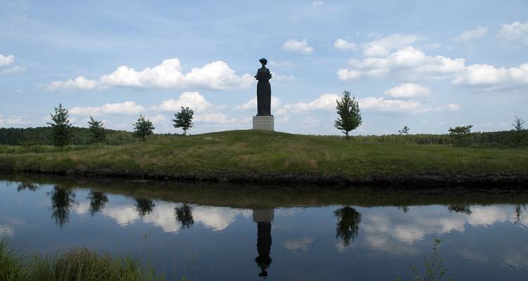 Eenzaam standbeeld bovenop een grasheuveltje weerspiegeld in een rustige gracht onder verspreide wolken.