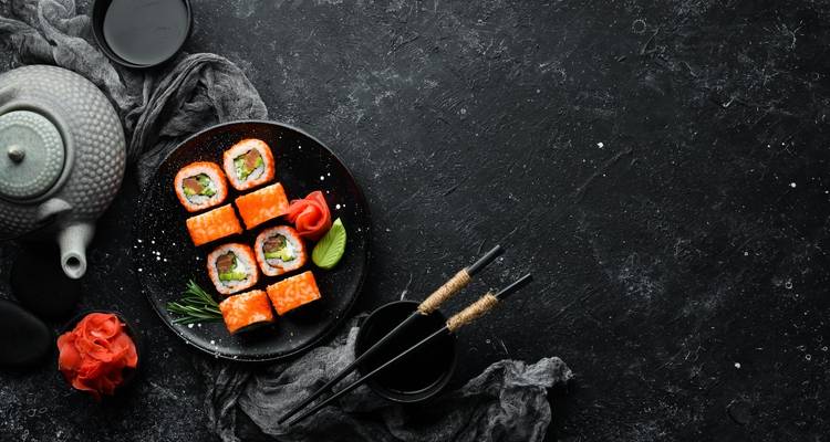 Draufsicht auf helle Sushi-Rollen mit Ingwer, Wasabi und Stäbchen auf einer dunklen strukturierten Oberfläche.