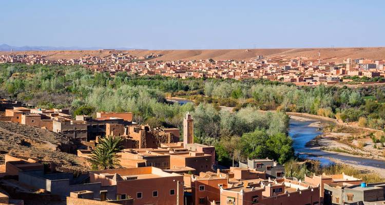Panoramic view of a Moroccan town with adobe buildings lining a winding river, lush green trees, and arid hills under a clear blue sky.