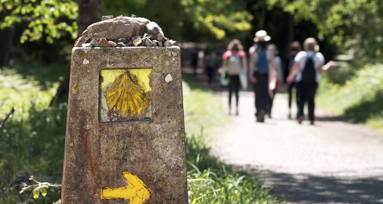 Marcador de piedra con un símbolo de concha de vieira y flecha amarilla, personas caminando por un sendero.