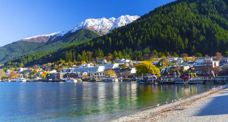 Sunlit Queenstown waterfront with alpine backdrop, forested slopes and clear lake waters.