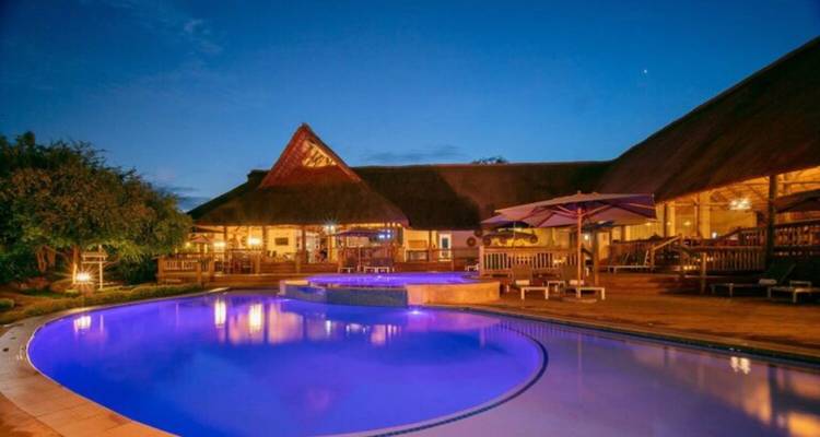 Hotelpool leuchtet blau in der Dämmerung neben Lodgegebäuden mit Strohdach.