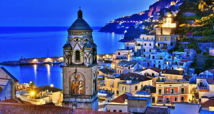 Vista al atardecer de la torre campanario de la catedral de Amalfi y las casas de colores pastel iluminadas contra un cielo azul profundo y el puerto.