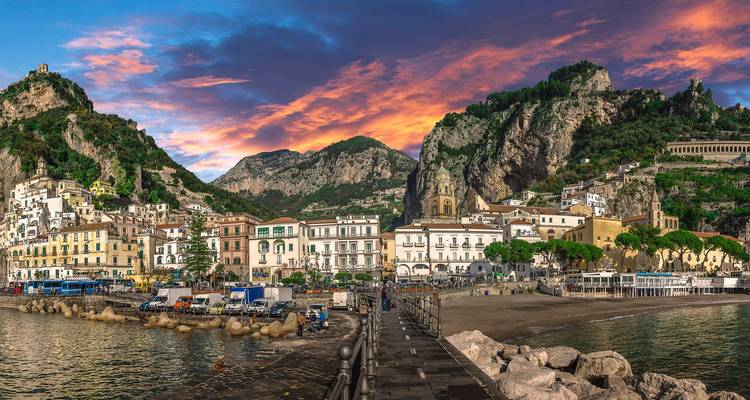 Panoramablick auf die Uferpromenade der Stadt Amalfi unter einem dramatischen rosa Sonnenuntergangshimmel, gesehen von einem steinernen Pier.