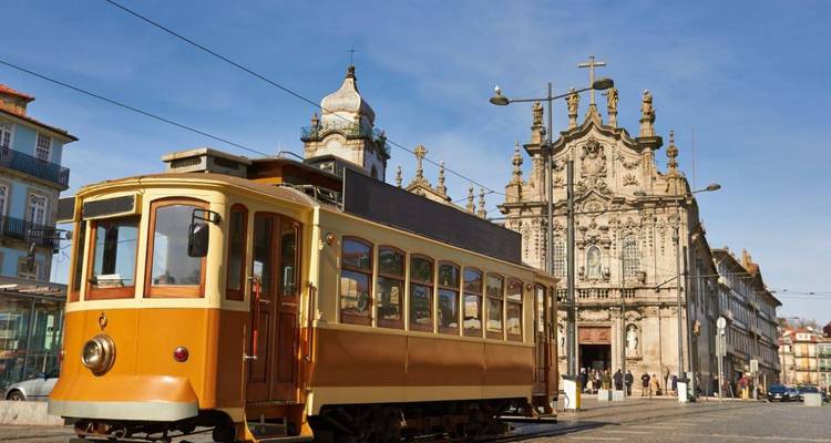 Eine historische gelbe Straßenbahn fährt an einer kunstvoll verzierten barocken Kirchenfassade in den Straßen von Porto vorbei.