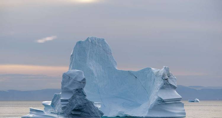 Majestic blue iceberg with sculpted peaks rises from calm Arctic waters under soft pastel sky.