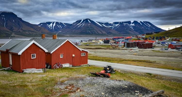 Rustic red cottages in the foreground overlooking fjord and serrated snowy mountains under heavy clouds