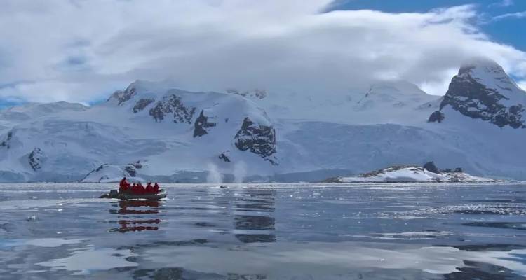 A Zodiac loaded with bundled explorers cruises icy Arctic waters beneath snowy peaks and clouds.