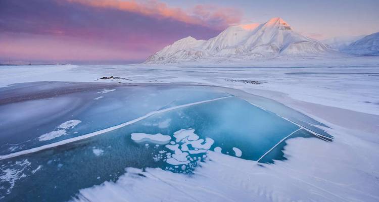 Frozen Arctic landscape with vivid turquoise ice pool, snow-covered mountains and pink twilight sky.