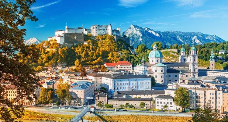 El horizonte de Salzburgo bañado en luz cálida con árboles teñidos de otoño, la catedral de cúpula y la Fortaleza de Hohensalzburg respaldadas por picos alpinos escarpados y un cielo azul despejado.