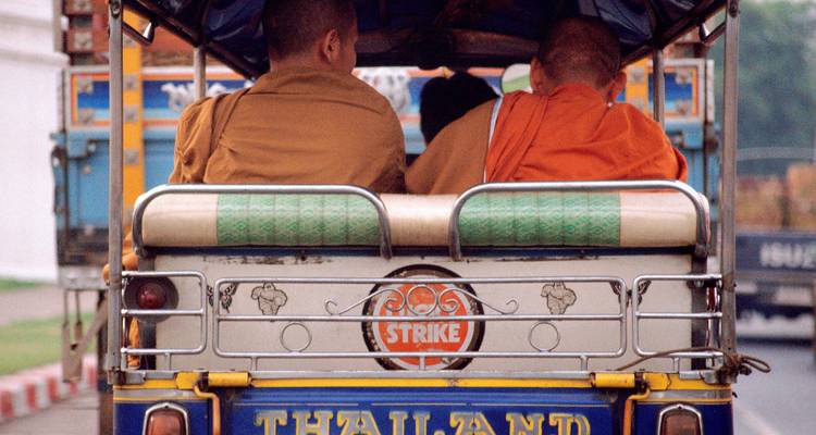 Dos monjes viajan en la parte trasera de un colorful tuk-tuk marcado con 'Thailand'.