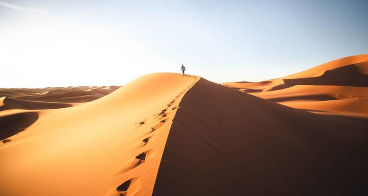 A lone figure walking along a sand dune at sunrise or sunset in the desert.