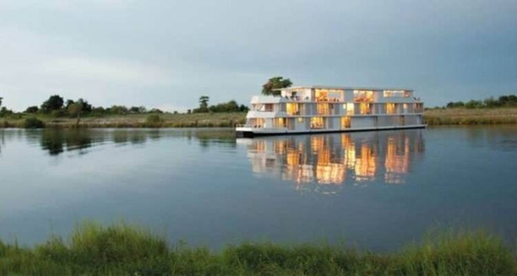 Modern luxury houseboat reflecting warm lights on calm river waters flanked by grassy banks at dusk.