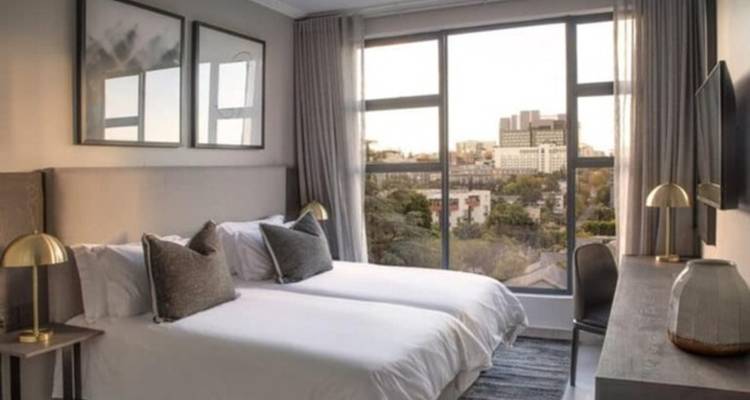 Contemporary twin-bed hotel room with large city-view window, neutral decor and soft evening light.