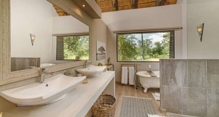 Spacious luxury lodge bathroom with double basins, standalone bathtub and woodland view through large window.