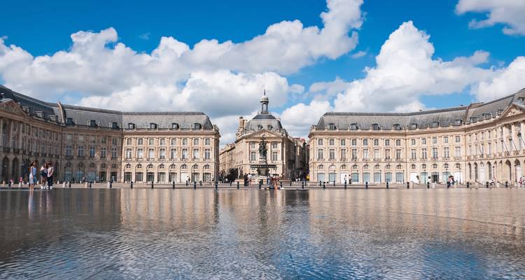 Symmetrischer Place de la Bourse spiegelt sich im Miroir d'Eau unter einem hellen, wolkengepunkteten Himmel wider.