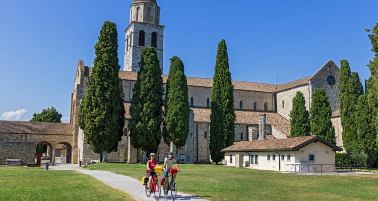 Cyclists ride past a historic abbey surrounded by tall cypress trees on a sunny day.