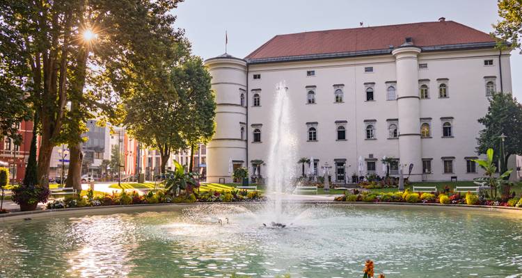 Elegant city square with a central fountain spraying water in front of a historic white building at golden hour.