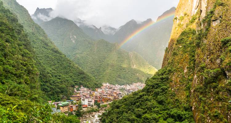 Luftaufnahme eines üppigen Tals mit einem Regenbogen und einer kleinen Stadt, die zwischen grünen Bergen eingebettet liegt.