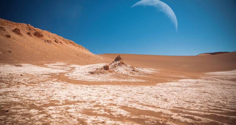 Lune au-dessus d'un paysage désertique austère avec du sable rougeâtre et des affleurements rocheux.