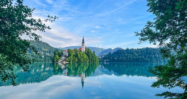 Mirror-like Lake Bled reflecting its island church and surrounding forested hills under a soft blue sky.
