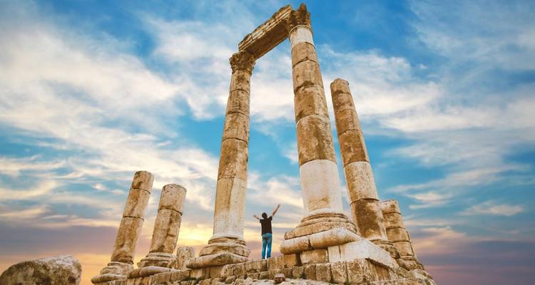 Person with raised arms stands amid towering ancient stone columns against a dramatic sunset sky.