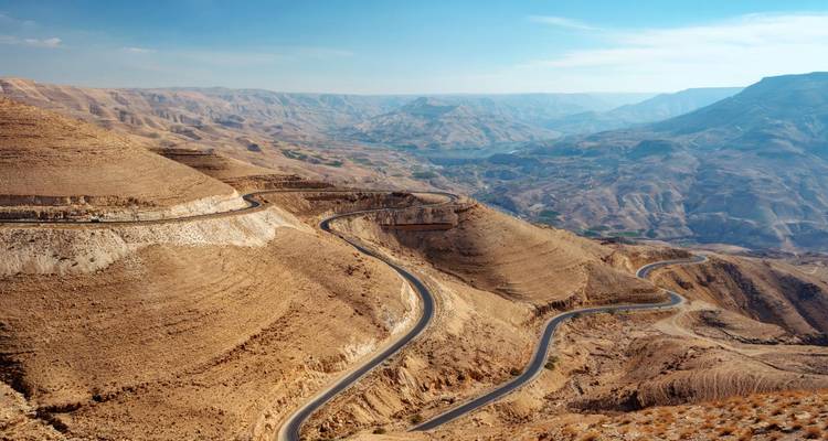 Snaking asphalt road winds through vast beige desert canyons and rugged hills under a blue sky.