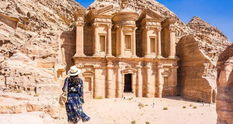 Woman in a floral dress and sunhat approaches the monumental rock-carved Monastery in Petra under clear skies.