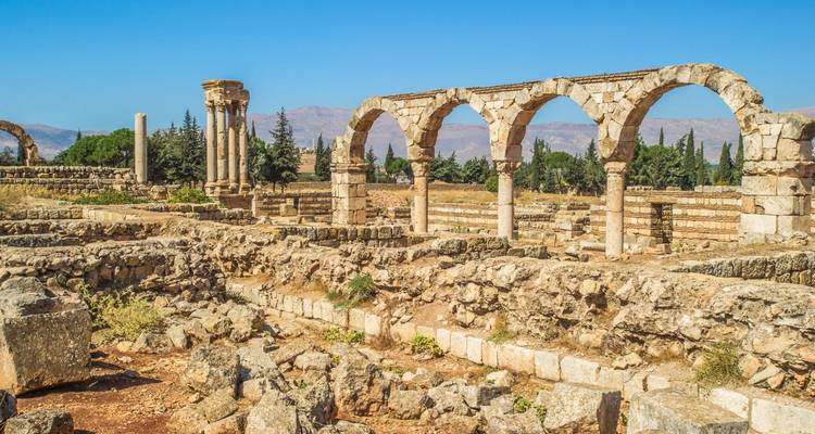 Sunlit remains of stone arches and columns stand amid sprawling ruins with distant cypress trees.