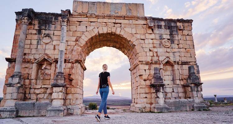 Voyageur marchant à travers une arche de pierre ancienne à Volubilis avec un ciel de coucher de soleil pastel.
