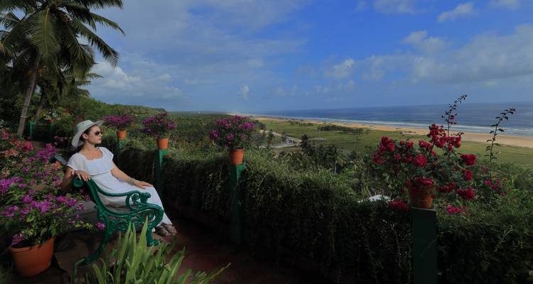 Femme en robe blanche assise sur une terrasse avec vue sur la mer et la plage.