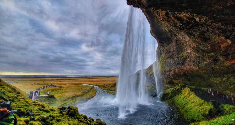 View from behind Seljalandsfoss waterfall cascading in front of a vast Icelandic plain under dramatic skies, with tiny visitors along the path.