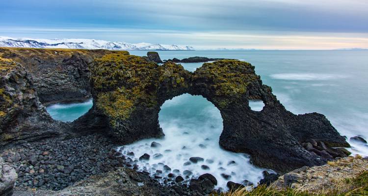 Basalt sea arch and rocky coastline at Arnarstapi with snow-capped mountains in the distance and smooth turquoise sea below.