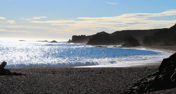 Sparkling sunlit waves crash on a black-pebble beach beneath rugged cliffs on the Snæfellsnes peninsula.