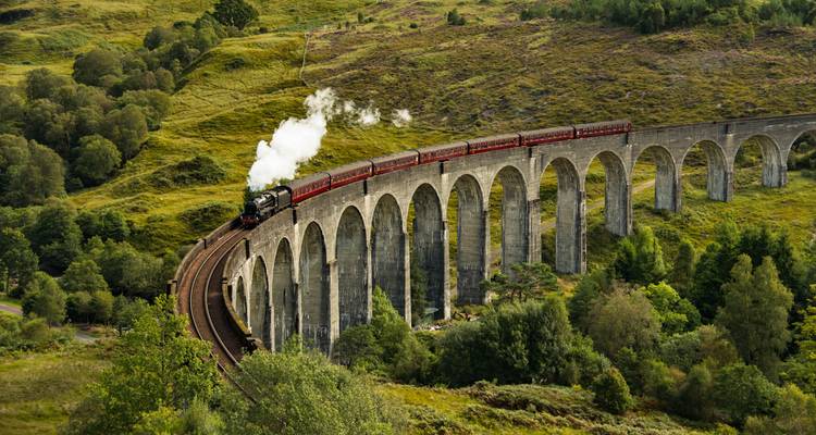 Steam train crossing a classic stone viaduct in a lush green landscape.