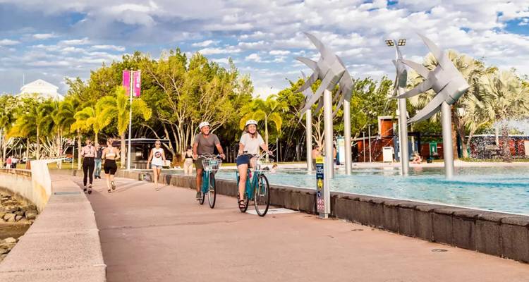 Des cyclistes sur un sentier près d'une fontaine et de sculptures, en plein air.