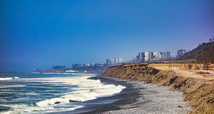 A rocky coastline with waves crashing and city skyline in the distance.