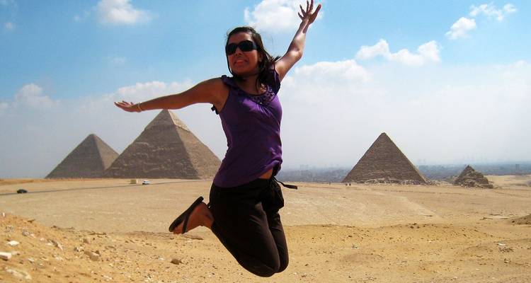Woman jumps mid-air in front of pyramids on the sandy Giza plateau.