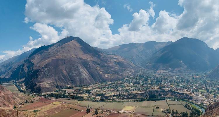 Panorama des Heiligen Tals von Peru mit Flickenteppich-Feldern unter türmenden Andengipfeln und flauschigen Wolken.